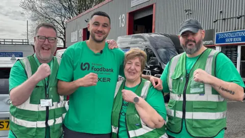 Four people stand in front of a large grey warehouse. They wear green branded t-shirts with "Barnsley Foodbank" written on them in black font. Callum Simpson, whose has short black, stands between a woman and two men who are all holding up a fist, imitating his boxer pose.