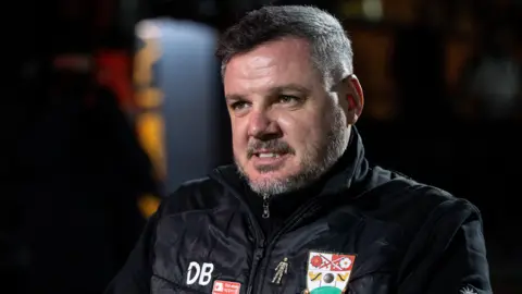 Dean Brennan wearing black a coat with the Barnet club badge and DB initials standing on the side of the pitch at full-time of a match