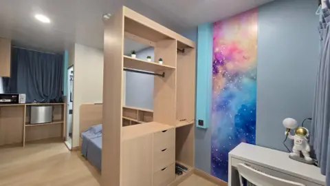 An empty room with a bed and shelf, as well as a small white table. 