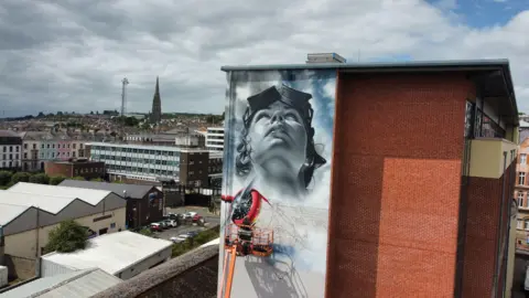 Michael Kielty Artist jeks stands on a cherry picker while paiting a mural of Amelia Earhart on the Foyle building in Derry, in the backgournd are the city's rooftops