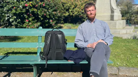 Liam Doherty pictured wearing a blue checked shirt and dark grey jeans sitting on a green park bench. He has dark brown short hair and a beard.