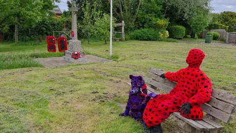 Kaz / BBC Weather Watchers A VE Day memorial featuring a figure covered in poppies sitting on a bench with a figure of a dog next to him. In the background there is a wreath in figure of 80 made from poppies next to a war memorial, with grass in the foreground