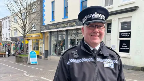 Inspector David Turnbull, a man wearing police uniform - including a flat hat - and glasses. He is standing in front of shopfronts in St Helier. A badge saying 'Police' is visible on is chest.