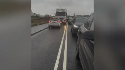 School Traffic is congested on a rural road. A building is to the side of the road. The sky is grey. It's been raining. 