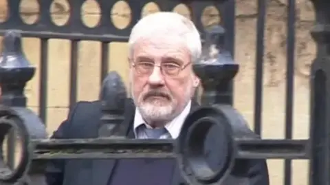 Stephen Cox, wearing a buttoned-up shirt and tie, but with the top button undone, and a purple jumper and dark jacket, walking out of Reading Crown Court, with the bars of railings in front of him as he walks down a ramp outside the building.
