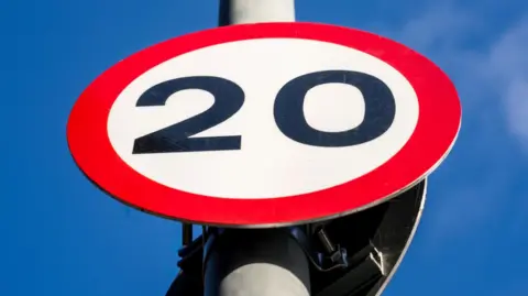 The picture has been taken looking up towards the round red and white sign with the number 20 in the centre of the sign. The sky is blue behind it.