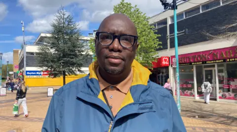 A bald man in a blue coat with yellow lining, wears a beige knitted sweater. He has dark-rimmed glasses and is standing in front of shops in Huyton.