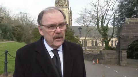 Elfyn Llwyd, standing outside Llandaff Cathedral wearing a black suit and tie with a white shirt and a black coat. He has a moustache and is balding with glasses 