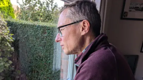 Andrew, a man with short gray hair and glasses stares out of his living room window.