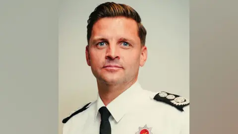 Hertfordshire Fire and Rescue Service A man with gelled ginger hair swept sideways. He is in a smart white shirt with black tie and the top of a red fire service badge on his chest. 