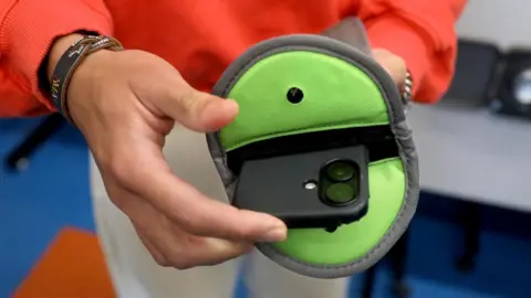 A school pupil wearing a bright orange jumper pushing a black phone into a green pouch.
