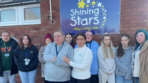 Nine women and one man stand outside a building. They are frowning and one woman in the centre has her arm around another woman. On the red brick building behind them is a blue and white sign which reads Shining Stars day care and club.