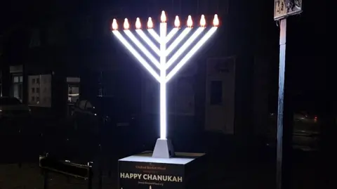 A street scene at night with a large menorah , which is the Jewish candelabra, with 9 bulbs on top. The stem and V-shaped prongs are lit up and glowing white. It stands on a black box with the words Chabad Bricket Wood wishes you a Happy Chanukah. Behind it are houses with cars on their driveways. 