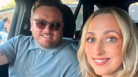 Family picture A selfie of Martyn and Gemma sitting in a car. They are both smiling. Martyn has brown hair and is wearing a blue zip up top with sunglasses. Gemma has long, curly blonde hair.