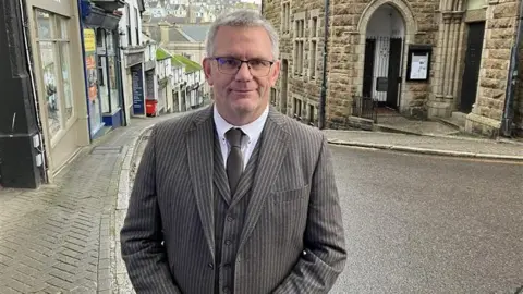 Johnnie Wells smiles as he stands on the pavement of the narrow road which has a sharp bend in it and leads down hill. He is wearing a brown pin striped three-piece suit with a brown tie. He has short grey hair and is wearing a pair of glasses.