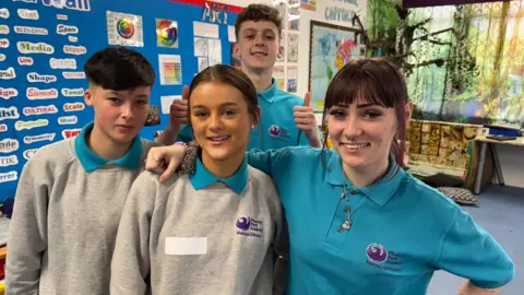 Four young students in one class. Two boys and two girls. One of the boys is parked behind the other three with both three thumbs, while a girl has scored her hand and goes to another. All four are posing for cameras.