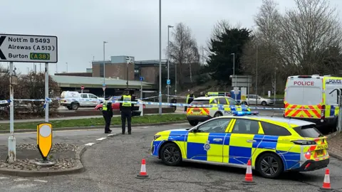 Police cars and a police van and cordon outside the station. At the front of the picture, a police car with blue and yellow markings is positioned and there are traffic cones on the ground. Four police officers are also in the picture. 