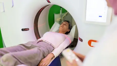 Stock photo shows a woman being inserted into an MRI machine with a person in the foreground holding a remote control, in a hospital environment.