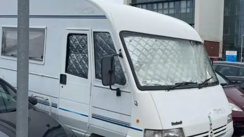 JO DUNNE The outside of the camper van, which is white and has foil weather protectors in the windows. It's parked between two cars with a large building in the background.