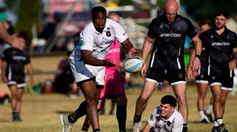 LooseHeadz A men's rugby sevens game taking place on a pitch. A man facing us, wearing a white kit, passes the ball towards us. A teammate is on the ground facing us. Two other players wearing black kits are standing near him. 