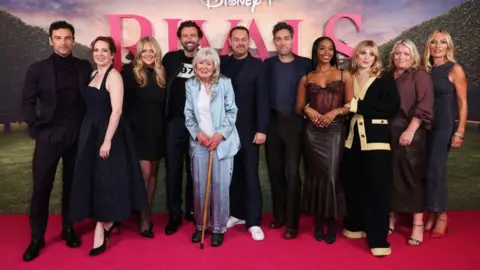Group image of actors who starred in Rivals. They are wearing evening dress and are smiling at the camera. They are standing on a red carpet.