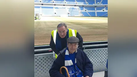 Gerry O'Brien/Everton FC John Gordon (front) wearing a grey cap and blue Everton scarf with a walking stick with Gerry O'Brien (back) standing behind him with hi-vis jacket on at Bramley-Moore Dock stadium.