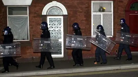 PA Media A single file of police in full riot gear and holding shields walk past terraced houses on a pavement in Oldham during a second night of disturbances on May 28, 2001.