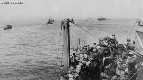 Black and white image of troops on a boat in the foreground on the right.  In the distance a number of other (some smaller) crafts, all heading towards the horizon.