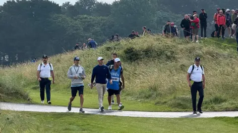 Men including a professional male golfer walk across a golf course carrying equipment. There are spectator standing up on a hill of grass watching them walk past.