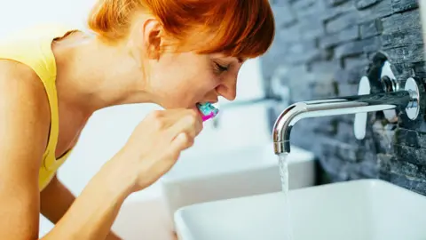 Getty Images Woman with red hair leans over a sink with the tap running to clean her teeth