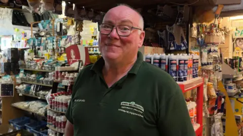 Paul Gillam is smiling at the camera inside his hardware shop. He is wearing a grey t-shirt and black framed glasses. The shelves are full of DIY supplies like paintbrushes, grout, paint and tape.