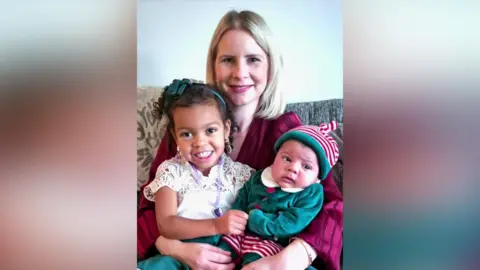 Becci Higgins, who is wearing a long-sleeved pink dress, is sitting on a patterned couch and cuddling her two children. 