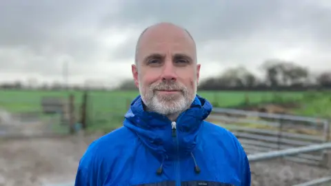 A man in a blue waterproof stands in front of a farm gate. There are green fields in the background.