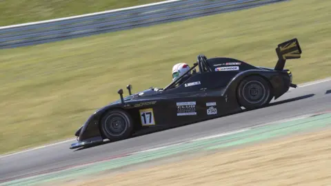 Ollie Read Photography Stephanie Hobeika wearing a helmet in a car, racing on a track at high speed. The car is branded and is a dark colour