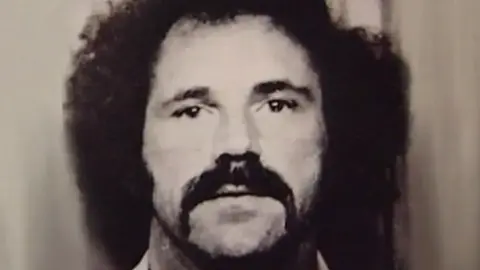 An old custody mugshot image of a young Ronald Evans. It is sepia and shows him with a long, dark handlebar moustache and mid-length hair. He has thick, dark eyebrows and is staring blankly into the camera.