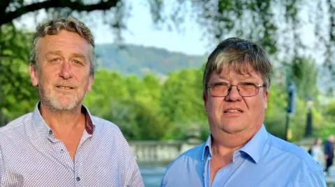 BANES Liberal Democrats David Harding on the left wearing a white and blue patterned shirt, standing beside Michael Auton on the right, who is wearing a blue collared shirt and glasses. They are both standing in front of a body of water with trees visible in the background.