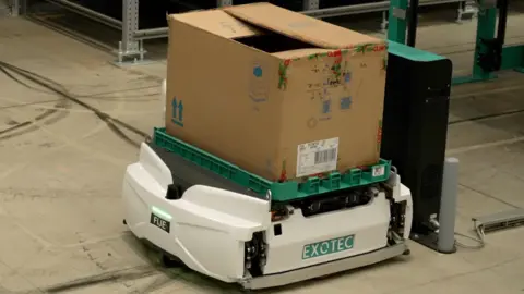 DJ McLaren/BBC A robot carrying a cardboard box, stationary on the floor of a warehouse. It has the letters "FIJE" on one side and the brand name "EXOTEC" on another. A green light shines above "FIJE". The box on top is open. There are black tyre tracks on the floor around the robot. 