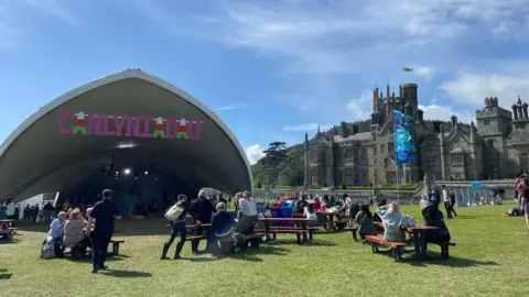 A covered stage sits on a field with a pink sign reading "Canlynliadau" meaning results in English. Margam Castle can be seen in the background on the right hand side. People sit on picnic benches in the sunshine outside the stage. 