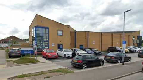 Google street view of Miranda House a two-storey building made from yellow brick with a large blue window at the corner and cars parked in front