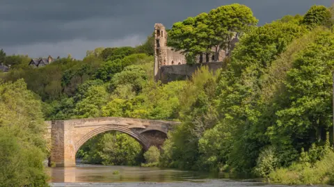 The ancient, ruined castle and its bridge across the River Tees. It is lit by warm, afternoon sunlight - but the sky is a deep, dark grey. Bright green trees with huge canopies of leaves partially obscure the castle and spread out from both banks of the river.