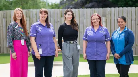 James Brew Photography Lady Peta Cavendish stands on green grass outside, in between to three hospice nurses in blue and purple tunics and Vicky Wilson who wears bright pink trousers and a colourful top.