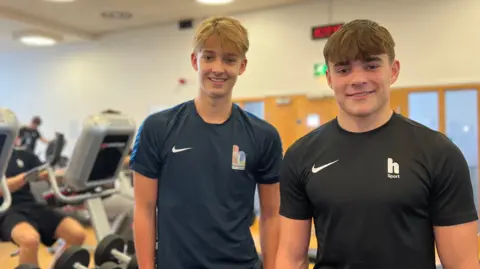 Oscar y Adam están parados cerca de un gimnasio con cintas de correr y mancuernas. Ambos visten ropa deportiva y camisetas con el logo de Nike. Al fondo se muestran otras personas utilizando máquinas cardiovasculares.