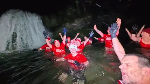 Group of people in bright red t-shirts and hats with black gloves in the water at the bottom of a waterfall.