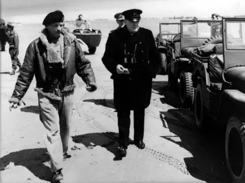 PA Media Sir Winston Churchill stands next to a row of military jeeps on a beach in Normandy. He is wearing a black overcoat, trousers and military hat. He is alongside Field Marshall Bernard Montgomery, who is wearing a beret and fur coat.
