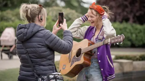 Run Away The character of Paige Greene, played by Ellie de Lange, is standing in Alexandra Park in Oldham with a guitar as a member of a woman, who is a member of the production company, takes a photograph of her using a mobile phone in front of a fountain.