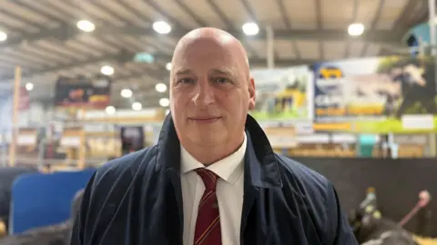 John McLenaghan, a man with a shaved head, looks to the camera while standing in a large, livestock shed at the Balmoral Show. He is wearing a white shirt, a maroon striped tie and a navy overcoat. 