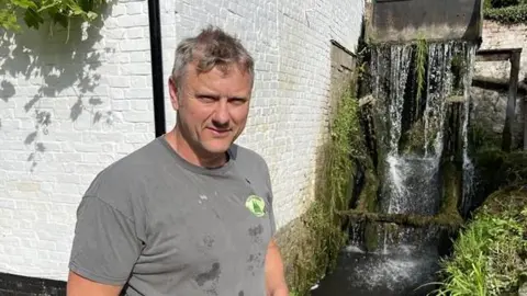 Phil Harrison / BBC A grey haired man in a khaki t-shirt stands in front of a white building and water mill.