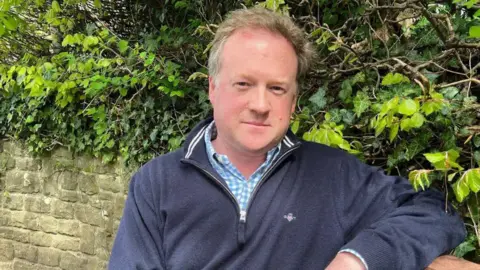 Close up image of man in blue shirt and blue jumper standing next to a hedge and wall