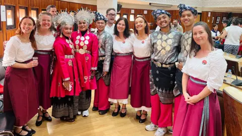 Skeddan Jiarg A group of Manx dancers in white tops and red skirts standing in a semi-circle alongside performers from other countries in their traditional dress, which is black, red and grey with gold discs and elaborate head dresses.