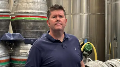 There is a man in a dark navy blue polo shirt. He has dark hair which is slightly sticking up. Over his left shoulder is a pallet of silver beer kegs, with a red and green line on. 
